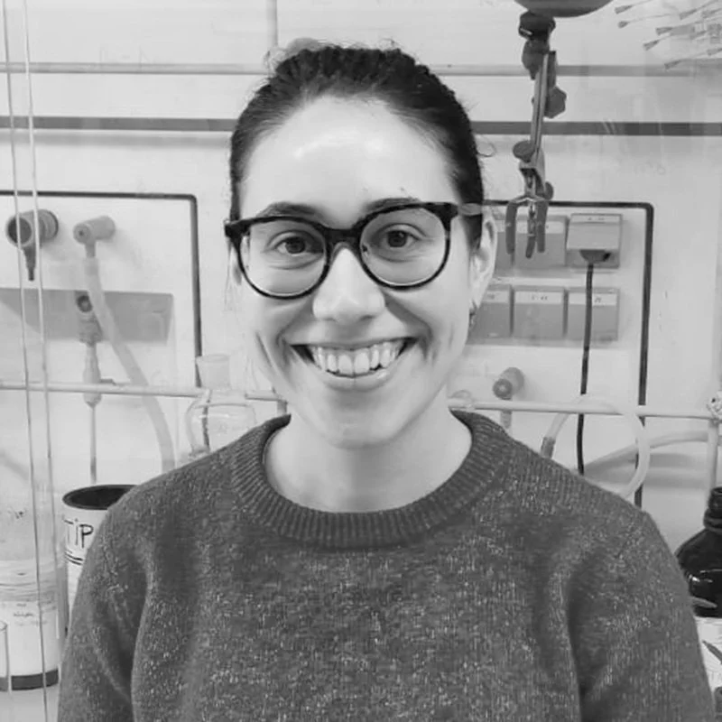 A woman smiling, wearing glasses and a sweater, in a laboratory setting with scientific equipment and apparatus behind her.