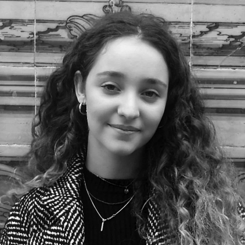 Black and white portrait of a young woman with curly hair, wearing a striped jacket, hoop earrings, and layered necklaces, sitting in front of wooden stairs.