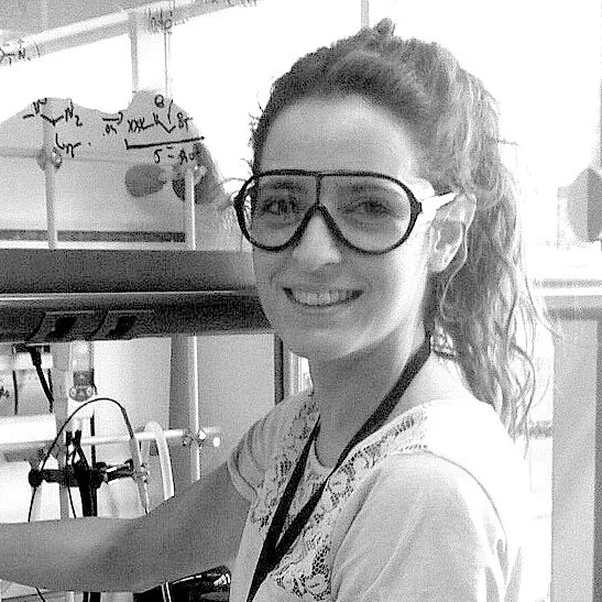Young woman in a science lab wearing safety goggles, smiling, with chemical formulas on the blackboard behind her.