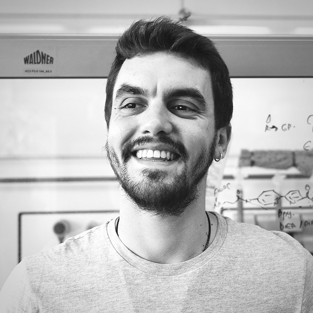 Black and white photo of a smiling man with a beard and earrings, standing in front of a whiteboard with scientific diagrams.