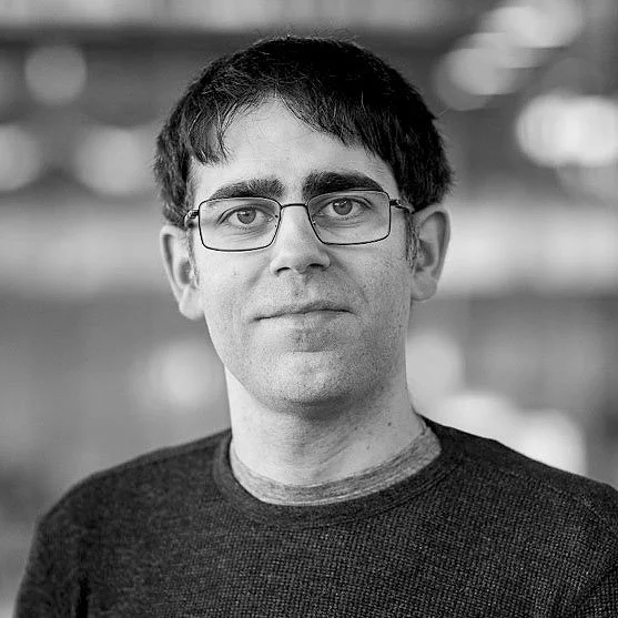 Black and white portrait of a man with glasses, short dark hair, wearing a dark sweater, looking at the camera with a neutral expression.