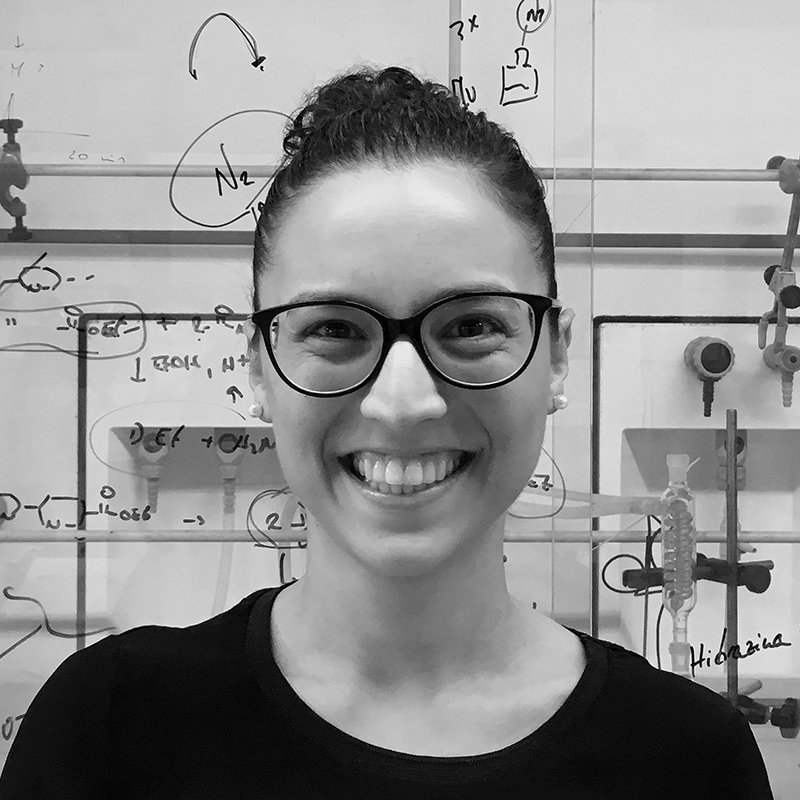 A woman with glasses smiling in a science laboratory with scientific equipment and handwritten notes on the whiteboard behind her.