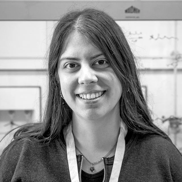 Black-and-white photo of a young woman with long hair, smiling, wearing a lanyard, in an indoor setting with a whiteboard or wall with writing in the background.