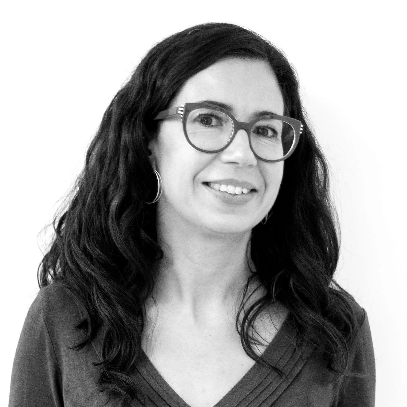 Black and white portrait of a woman with long curly hair, wearing glasses and hoop earrings, smiling slightly, against a plain light background.