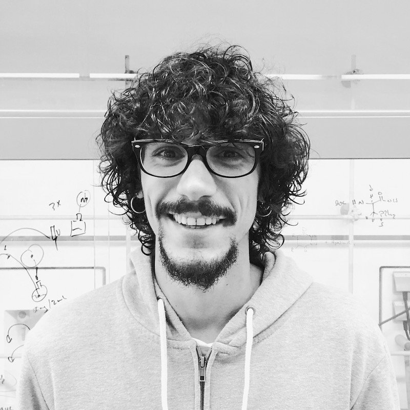 A young man with curly hair, glasses, and a beard smiling at the camera, wearing a hoodie, standing in front of a whiteboard with technical diagrams and writings.