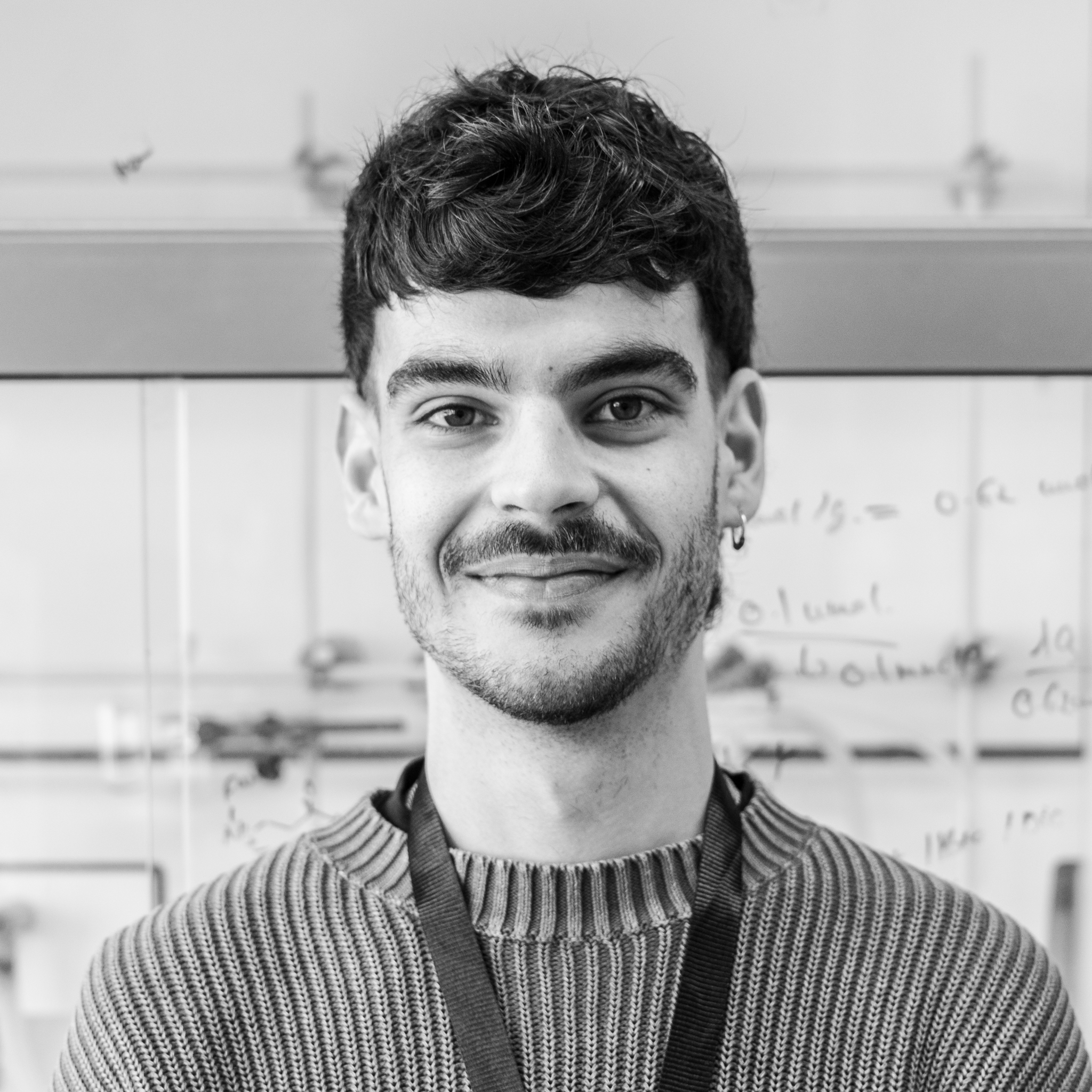 A young man with dark, wavy hair, a goatee, and an earring, smiling at the camera in a classroom or lab environment, with whiteboards and scientific equipment in the background.