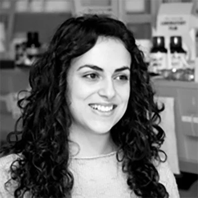 A woman with curly hair smiling in what appears to be a store or pharmacy setting.