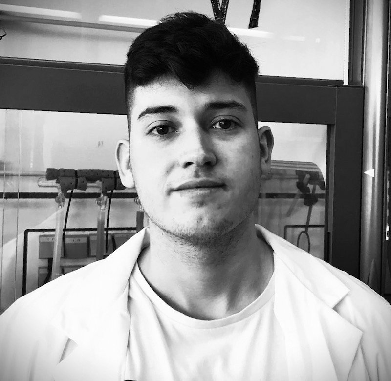 A young man with short dark hair, wearing a white lab coat, standing indoors in front of laboratory equipment.
