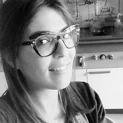 A woman with medium-length hair wearing large glasses and a dark top, smiling at the camera in a kitchen setting.