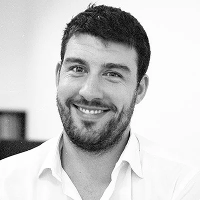 A smiling man with dark hair and a beard, wearing a white shirt, in a black-and-white photo.