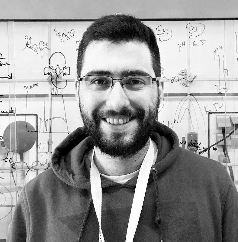 Young man with glasses and a beard smiling in front of a whiteboard filled with scientific equations and diagrams.