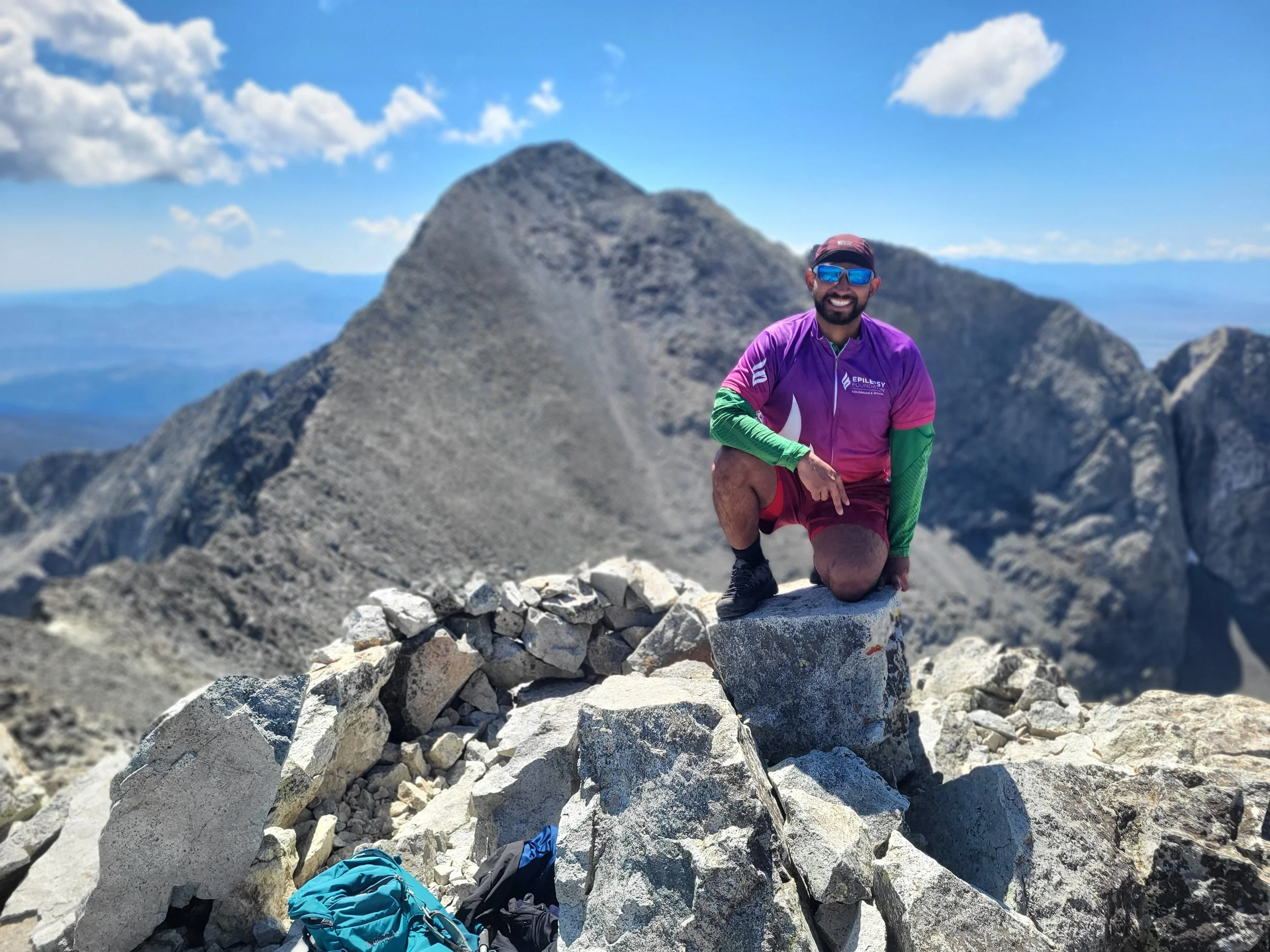 Sal at the summit of a 14er smiling, wearing a purple Epilepsy Foundation shirt, sunglasses, and a cap, with a mountain landscape and blue sky in the background.