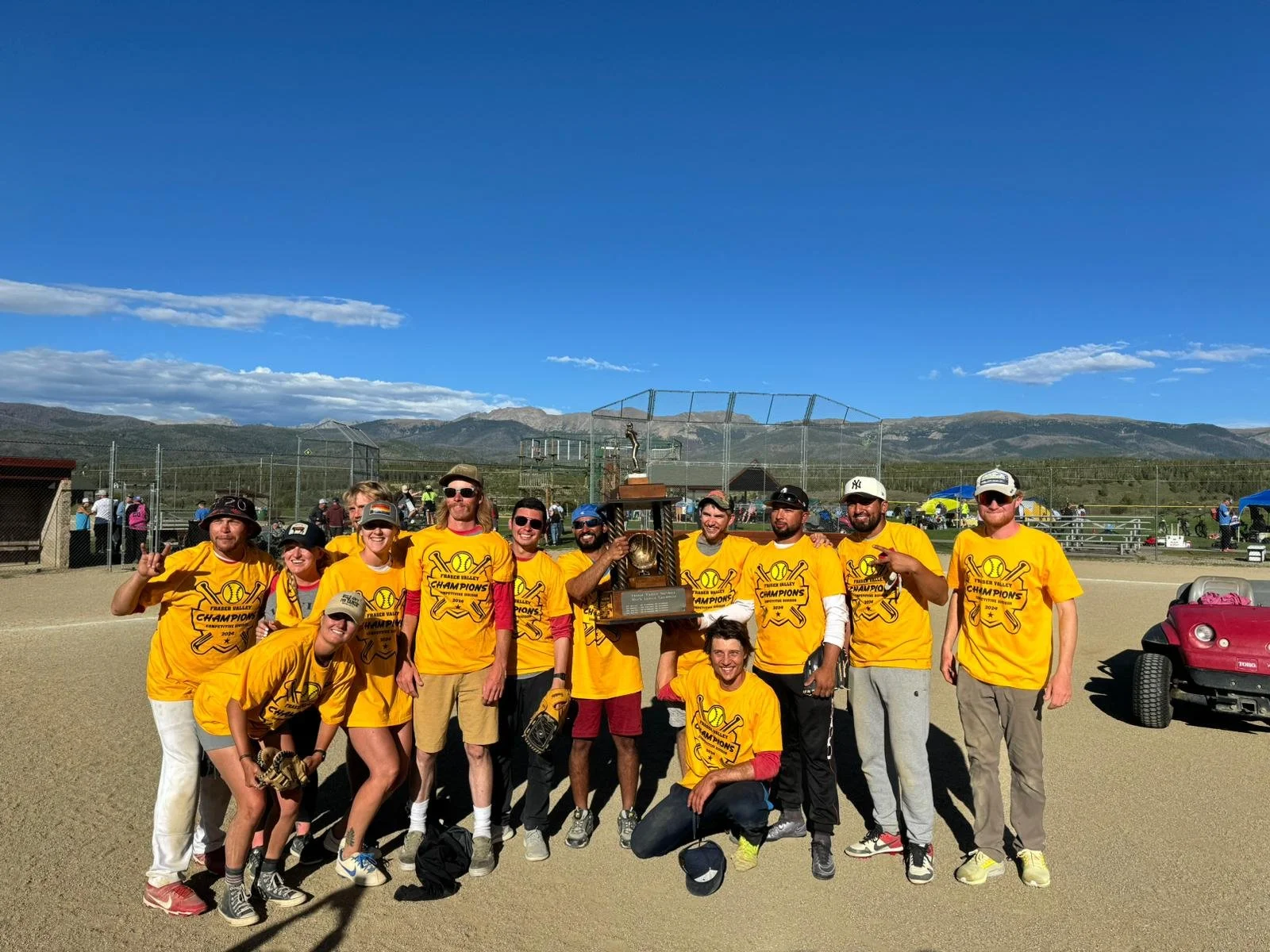 Sals team, celebrating winning the Fraser Valley rec devision. They are holding a large trophy, smiling, and some are making victory signs. The background shows a softball field, fencing, and mountains under a blue sky.