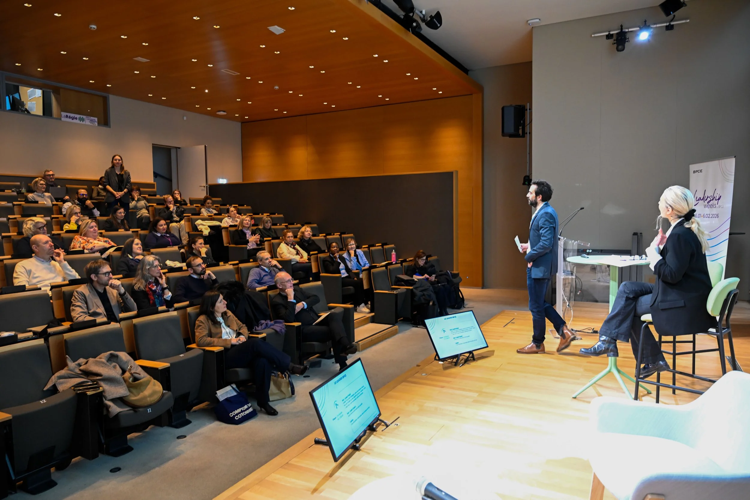 Conférence dans une salle avec deux intervenants se tenant devant un public assis. L'un des intervenants, un homme en costume, parle, tandis qu'une femme en veste noire écoute.
