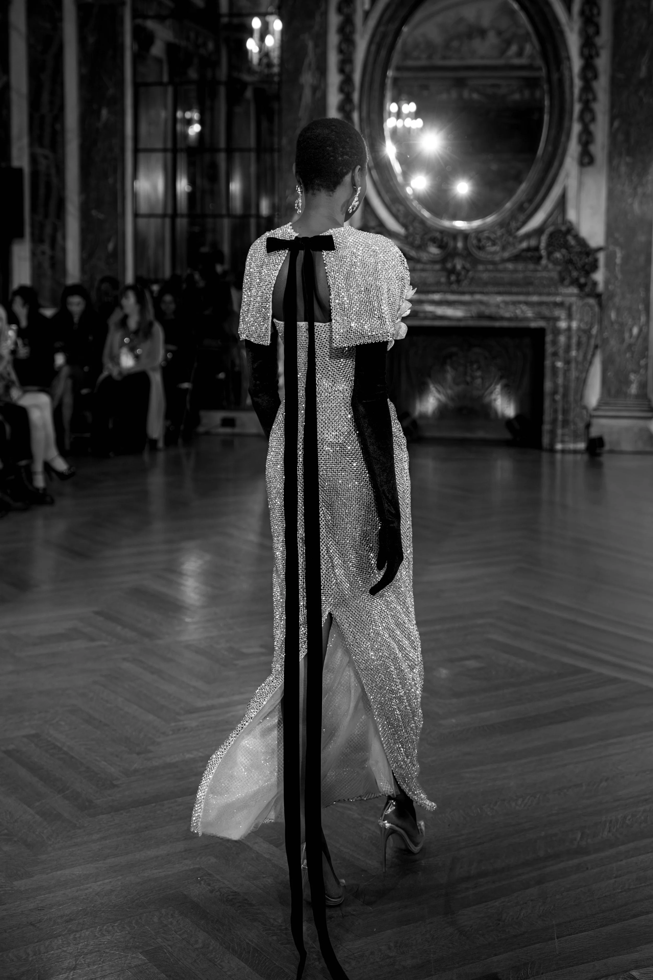 A woman on a runway in an elegant, sparkling evening gown with a black ribbon detail, in a grand, ornate room with a fireplace and mirror.