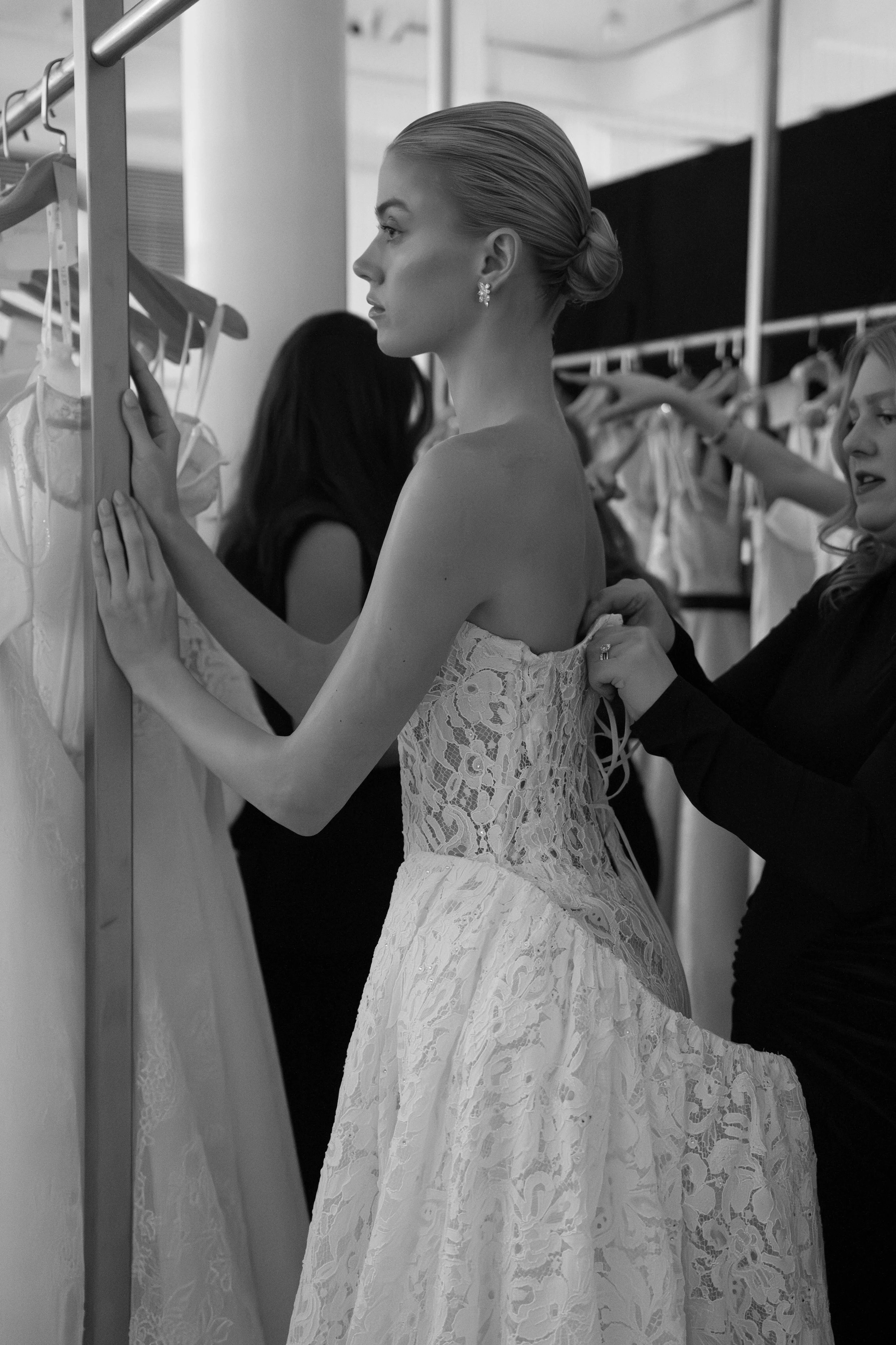 A woman in a strapless lace wedding dress trying it on while another woman helps adjust the dress in a bridal boutique.
