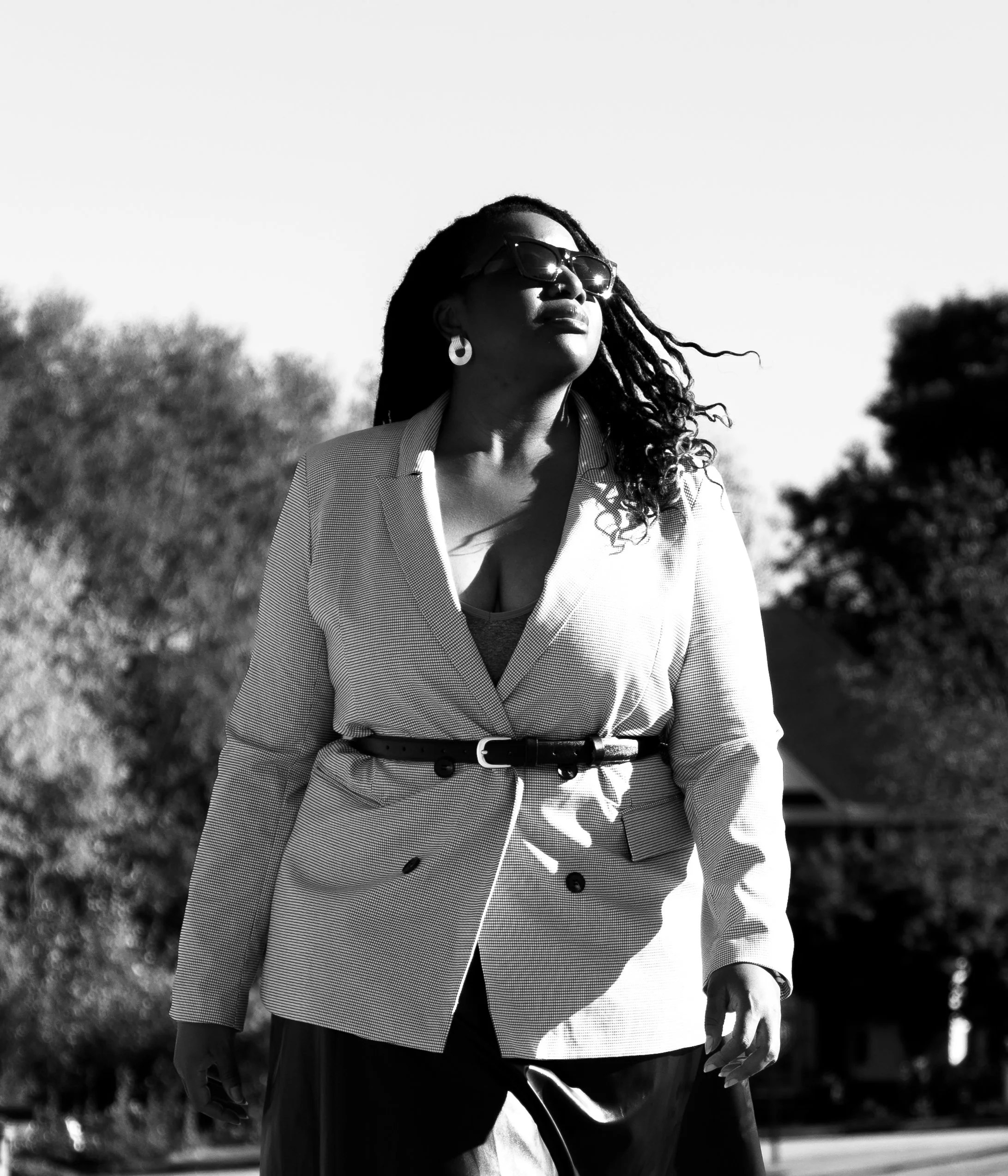 A woman with dreadlocks, sunglasses, and earrings, dressed in a blazer and top, walking outdoors in sunlight.