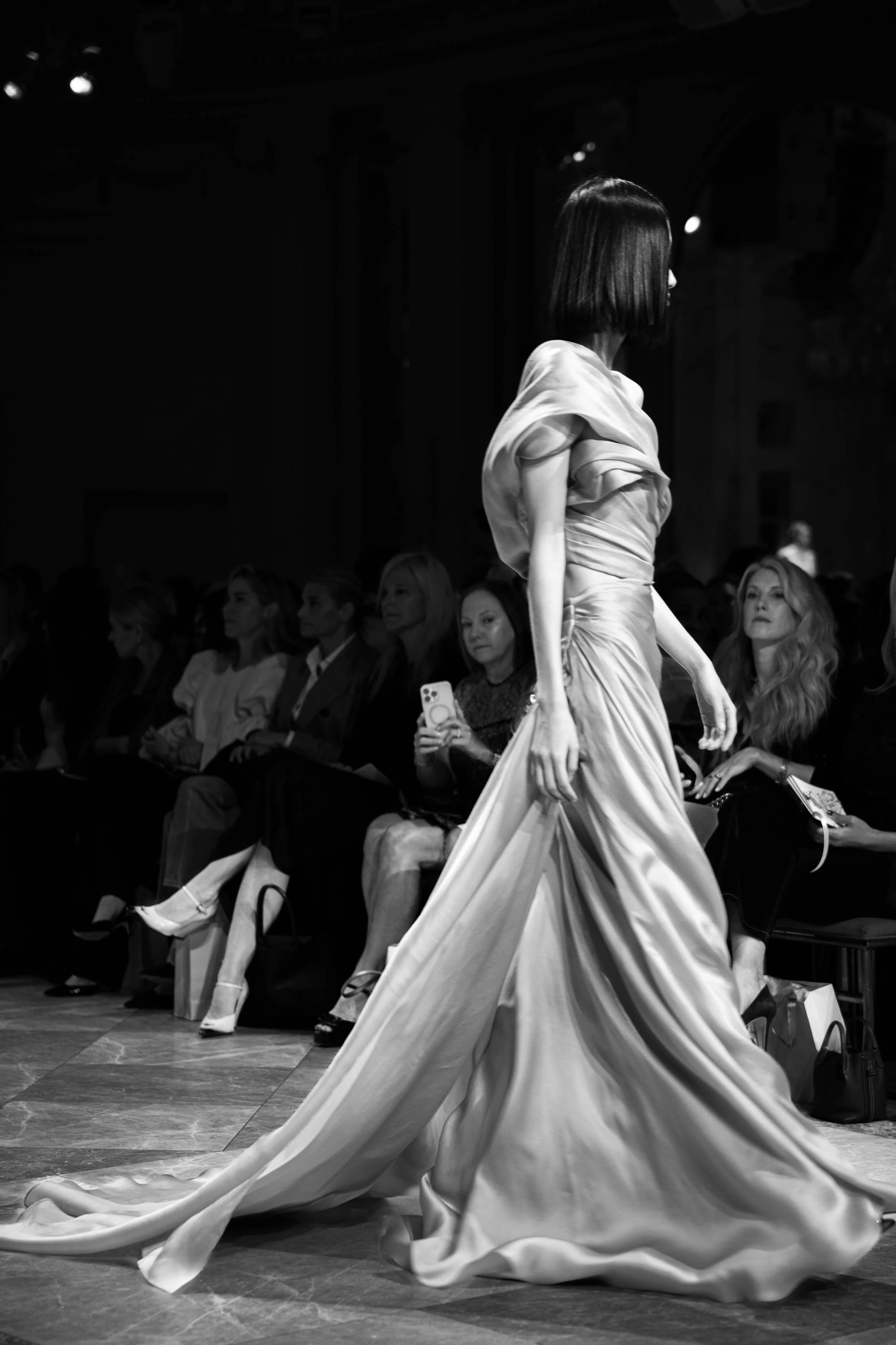 Black and white photo of a model walking on a runway in a couture gown, with seated spectators in the background at a fashion show.