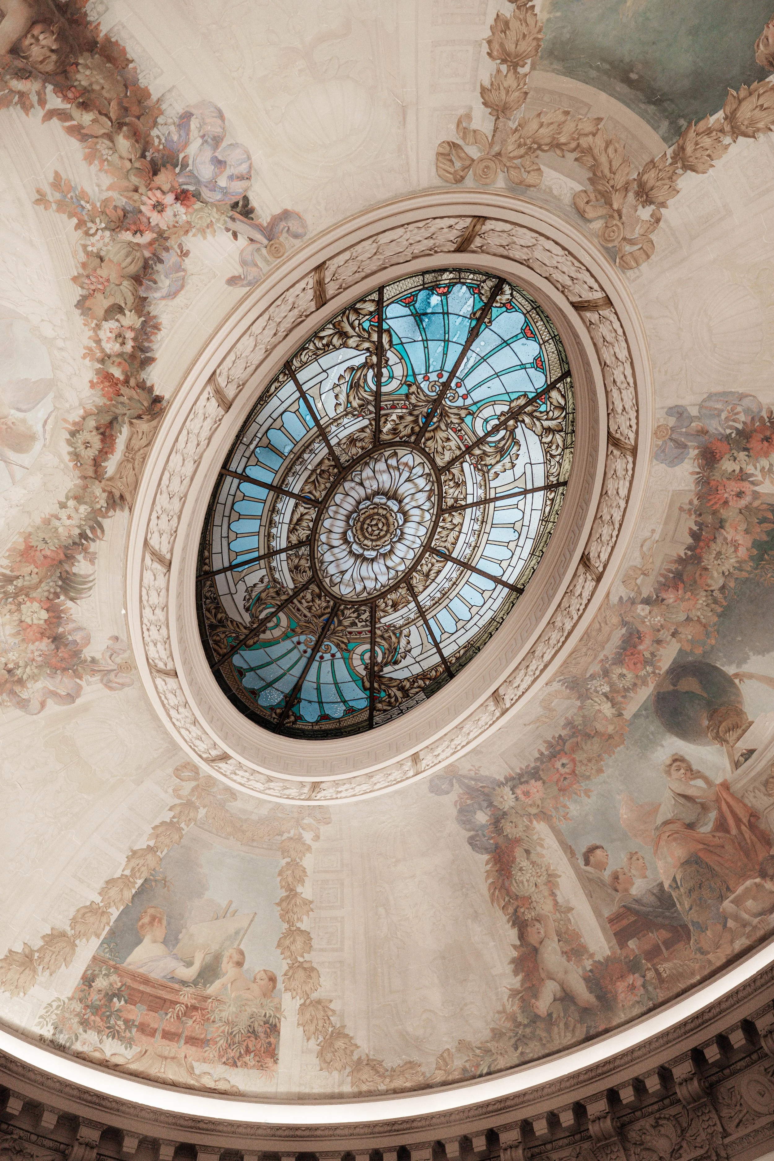 Interior view of a domed ceiling with a stained-glass skylight, decorated with intricate floral and ornamental artwork, and paintings of people.