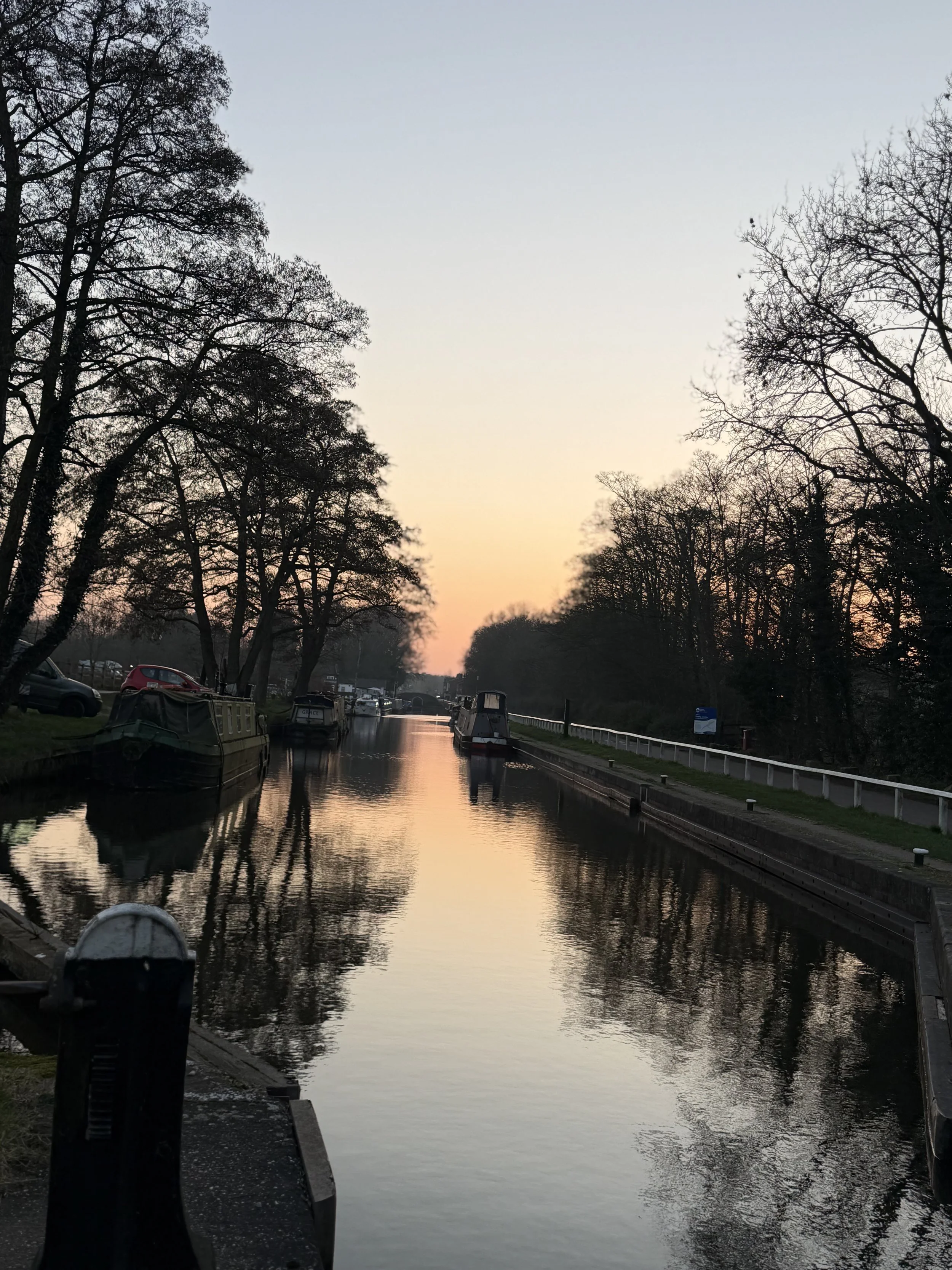 Calm canal water reflects trees and boat silhouettes at sunset, with a pathway and parked boats on the sides.
