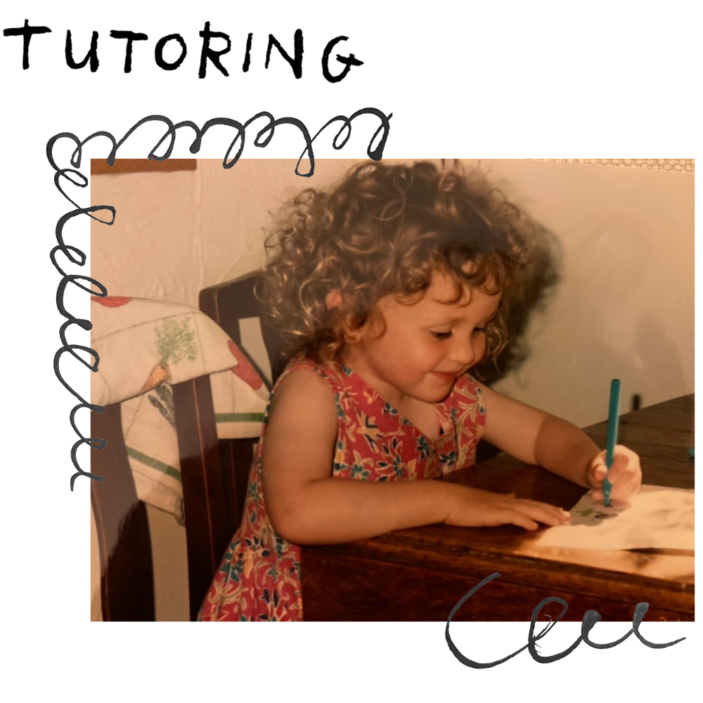 A young girl with curly hair sitting at a table, drawing on a piece of paper with a blue marker.