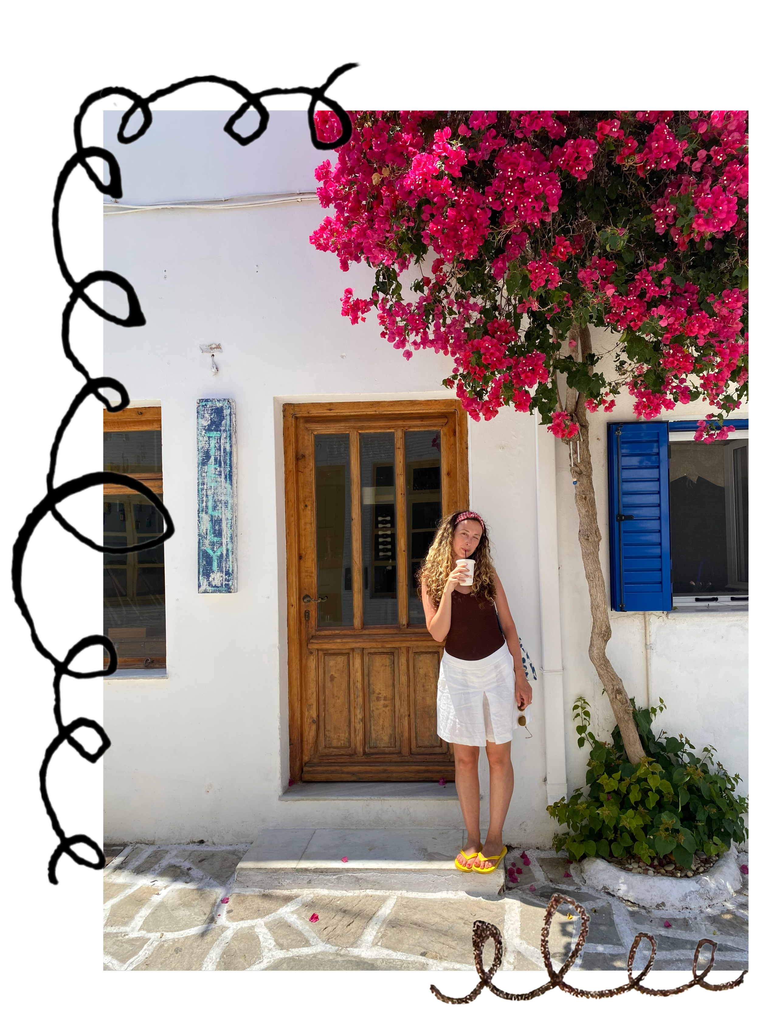 A young woman with curly hair wearing a headband, black sleeveless top, and white skirt, stands outside a white building with a wooden door, blue window shutters, and a pink flowering tree, holding a cup close to her mouth.