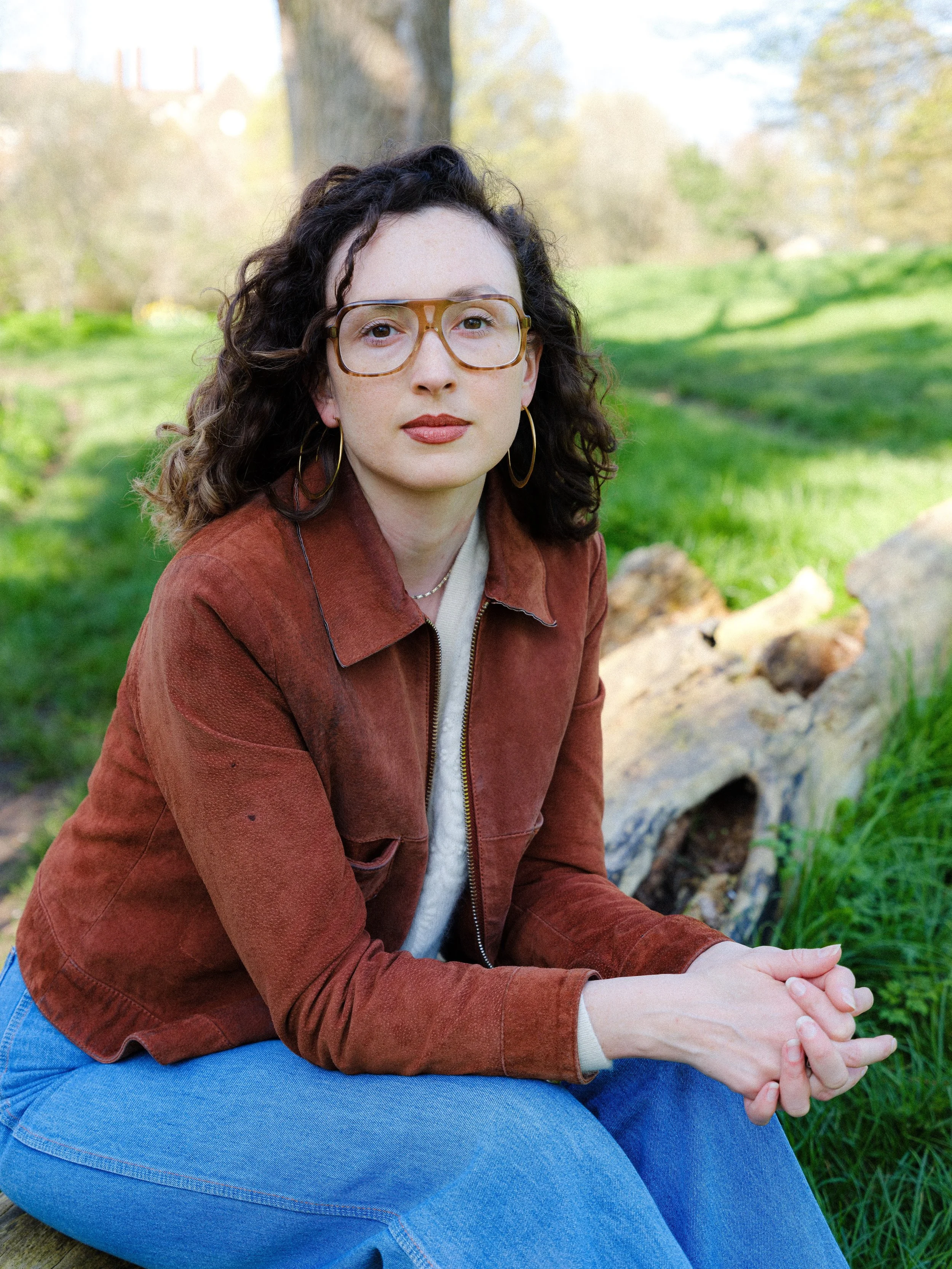 A woman with curly dark hair, wearing large glasses, a brown suede jacket, and gold hoop earrings, sitting outdoors on a green grassy area with rays of sunlight and trees in the background.
