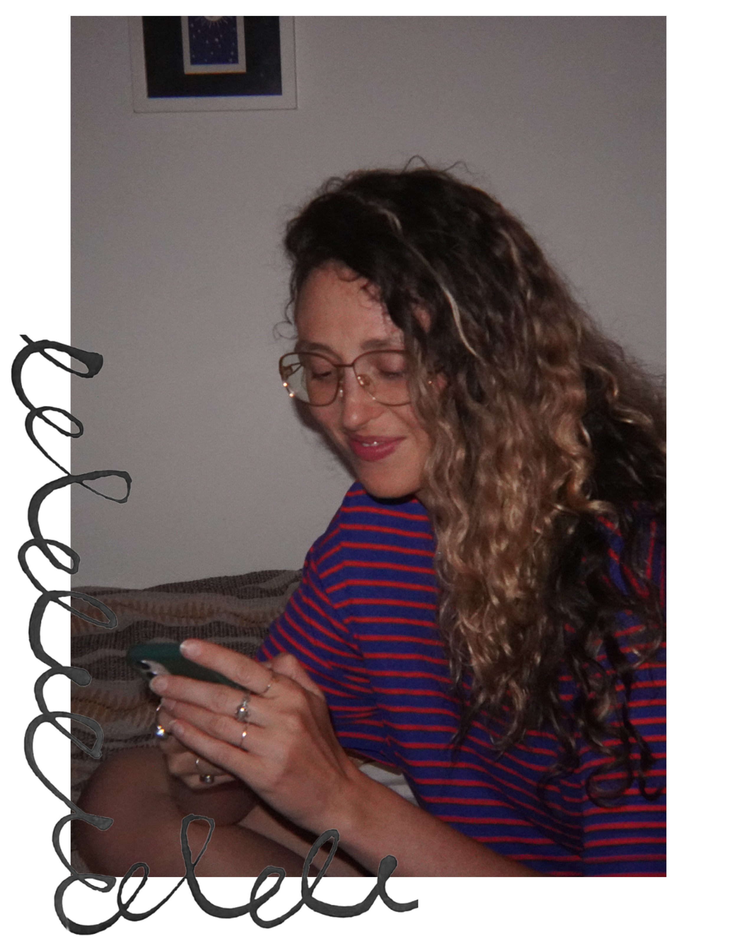 A young woman with curly hair and glasses, wearing a red and blue striped shirt, sitting on a bed and looking at her phone.