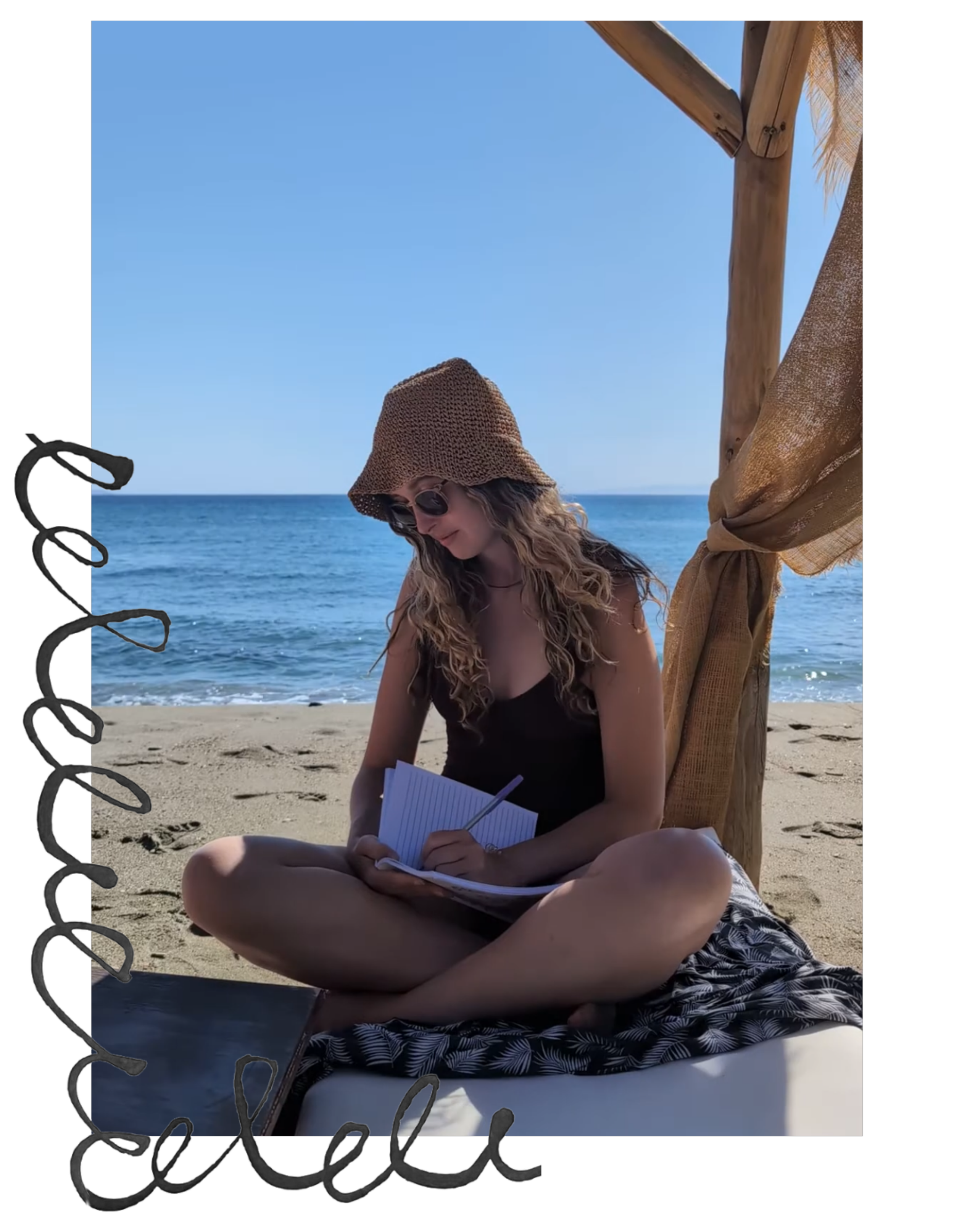 A woman sitting cross-legged on a beach mat, writing in a notebook, with the ocean and clear blue sky in the background, wearing a brown sunhat and sunglasses.