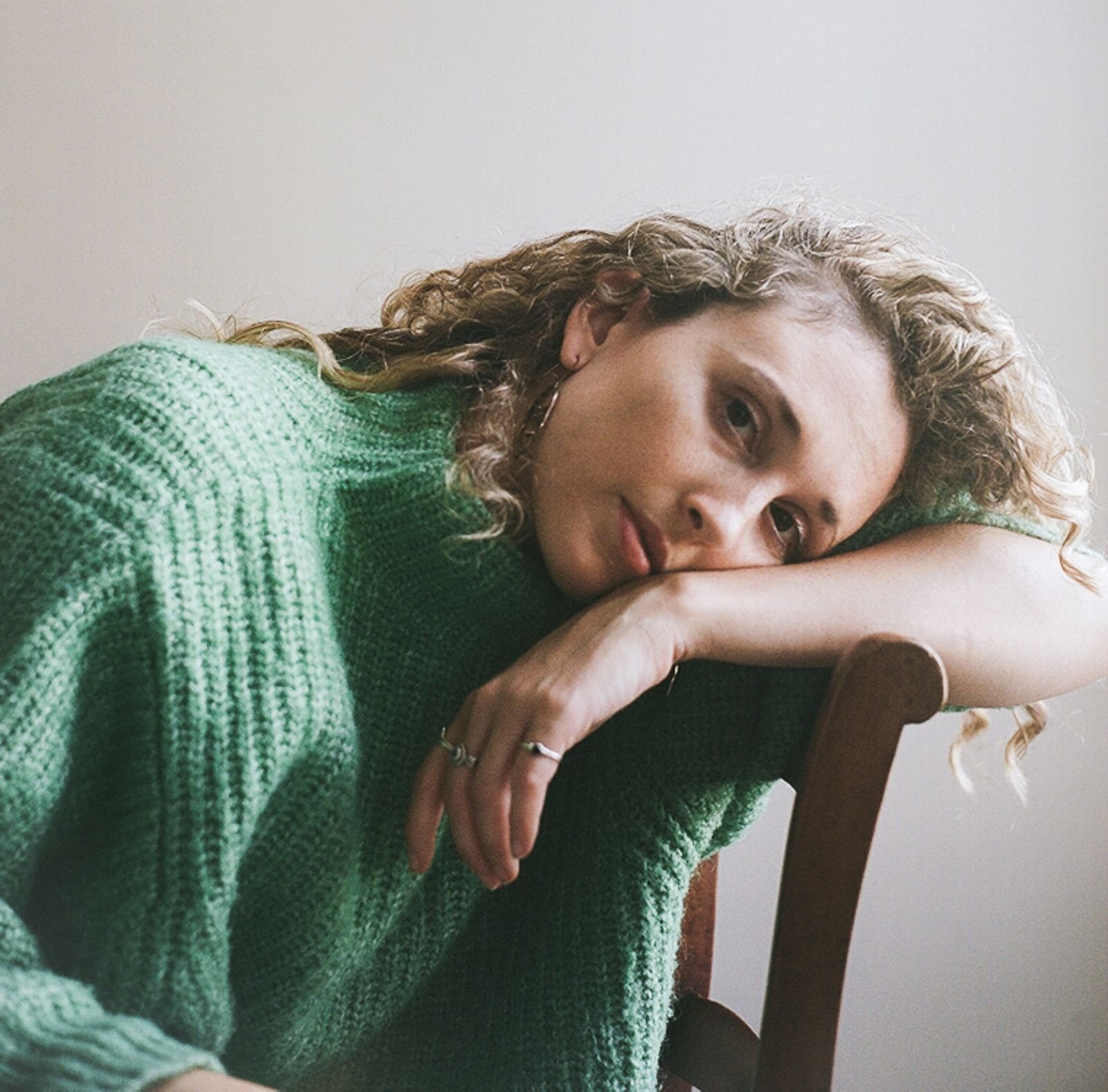 A woman with curly hair in a green knitted sweater resting her head on her crossed arm on the back of a chair.
