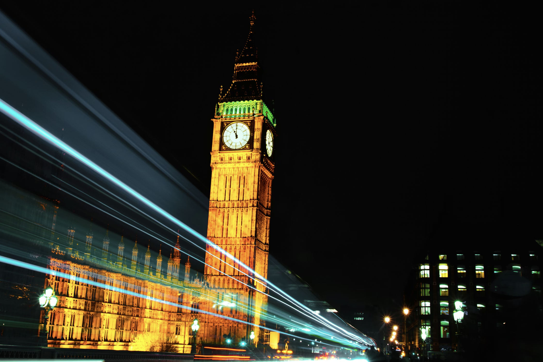 Nighttime photo of Big Ben in London illuminated with orange and green lights, with light trails from moving vehicles in the foreground and a dark sky in the background.