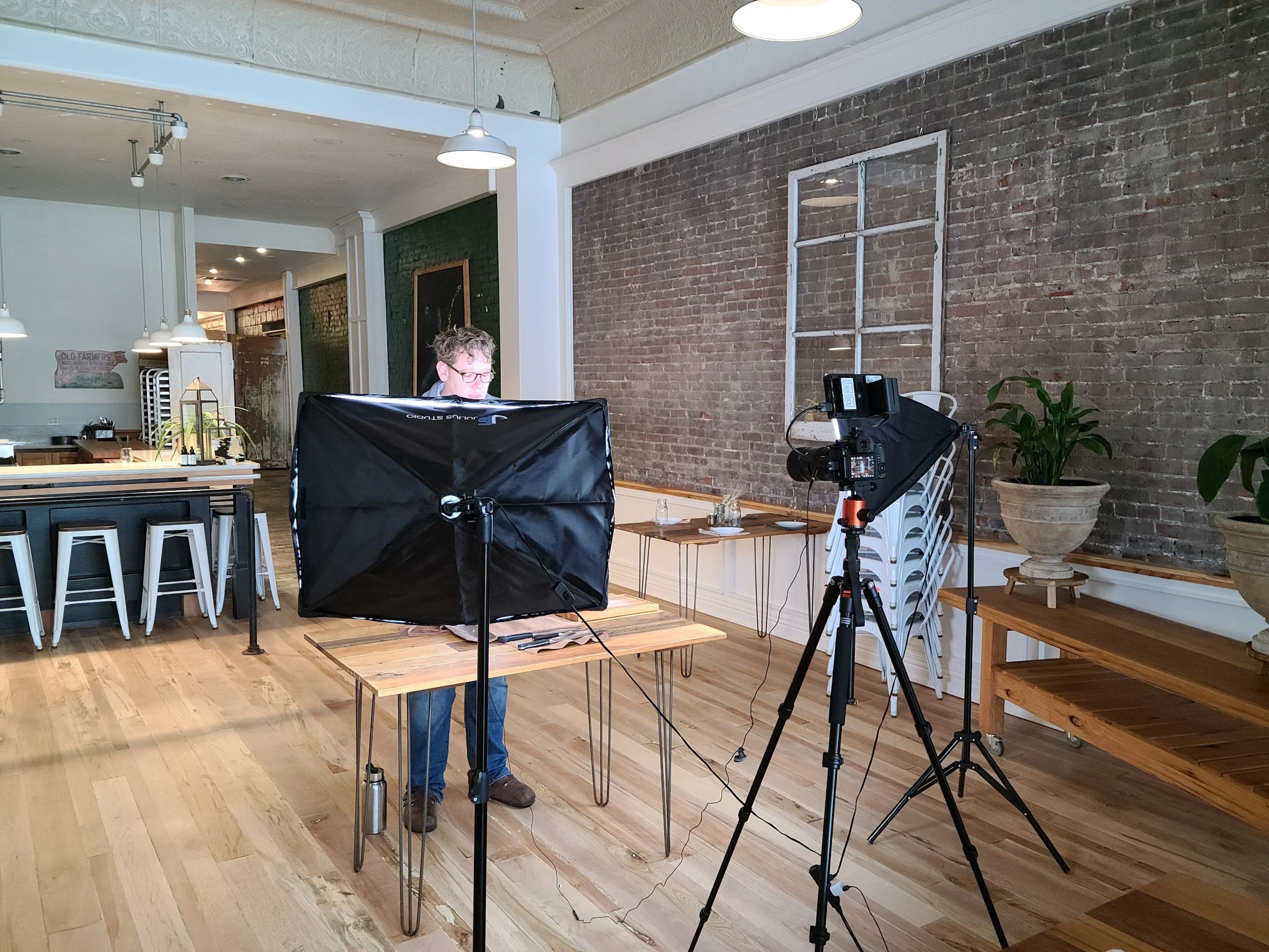 A man setting up a video shoot in a cafe with professional lighting and camera equipment, wooden floors, brick wall, and potted plants.