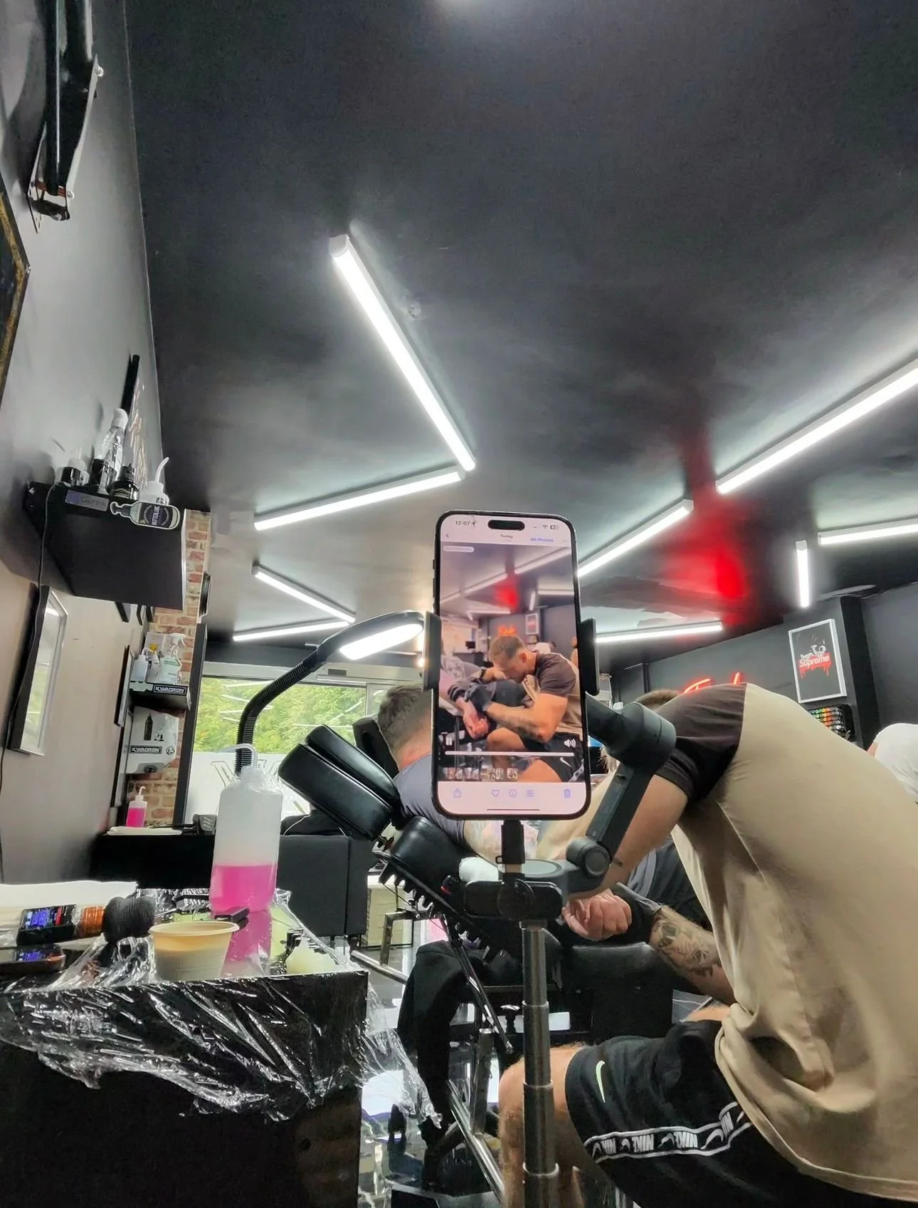 Tattoo artist tattooing a person's arm while another person is recording with a smartphone in a tattoo studio.