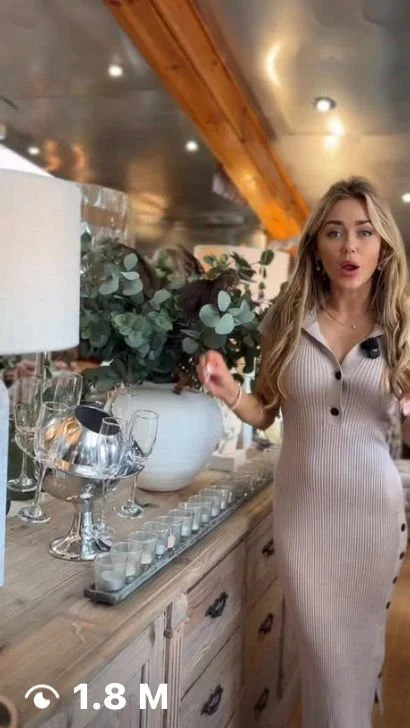 A woman with long blonde hair wearing a beige ribbed dress with buttons, standing next to a wooden sideboard with decorative items and a large bouquet of eucalyptus and greenery in a white vase. She is indoors with a warm, rustic ceiling.