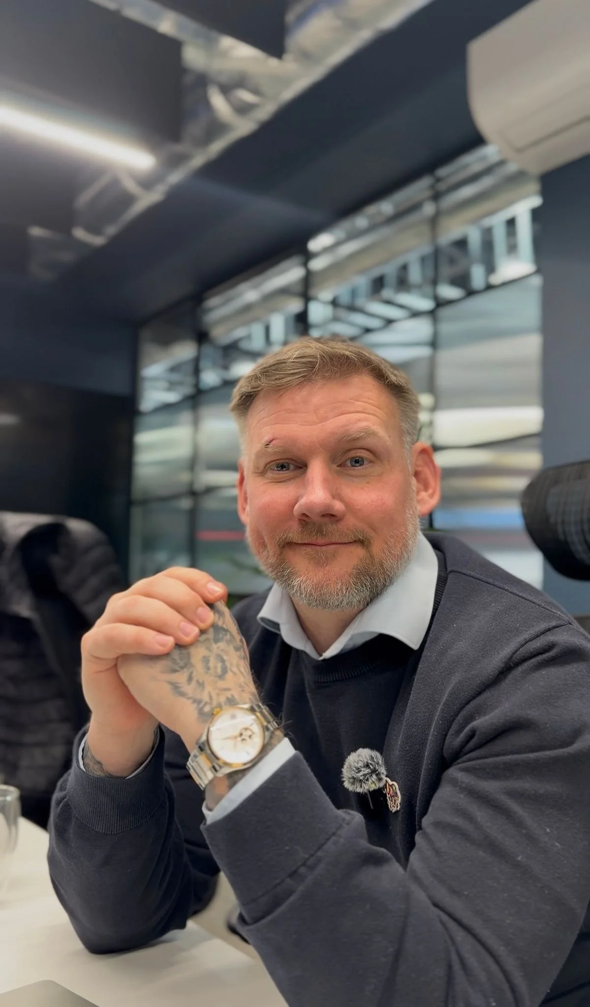 A man with a beard and tattoos on his hand, wearing a watch, sits at a table with his hands clasped. He is smiling at the camera in an indoor setting with large windows and modern decor.