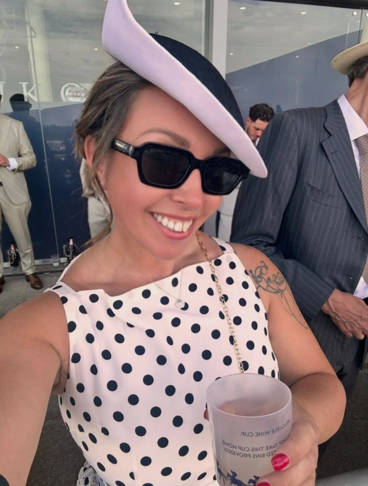 A woman smiling, wearing a white dress with black polka dots, large black sunglasses, a pink and white hat, and holding a clear plastic cup with text. She has a tattoo on her left shoulder and is at a social event with other people in the background.