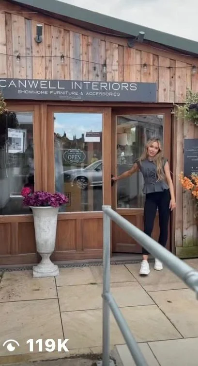 A woman standing outside Canwell Interiors, a showhome furniture and accessories store, on a sidewalk with a large flowerpot, wooden storefront, and glass door. She is smiling and holding the door handle.