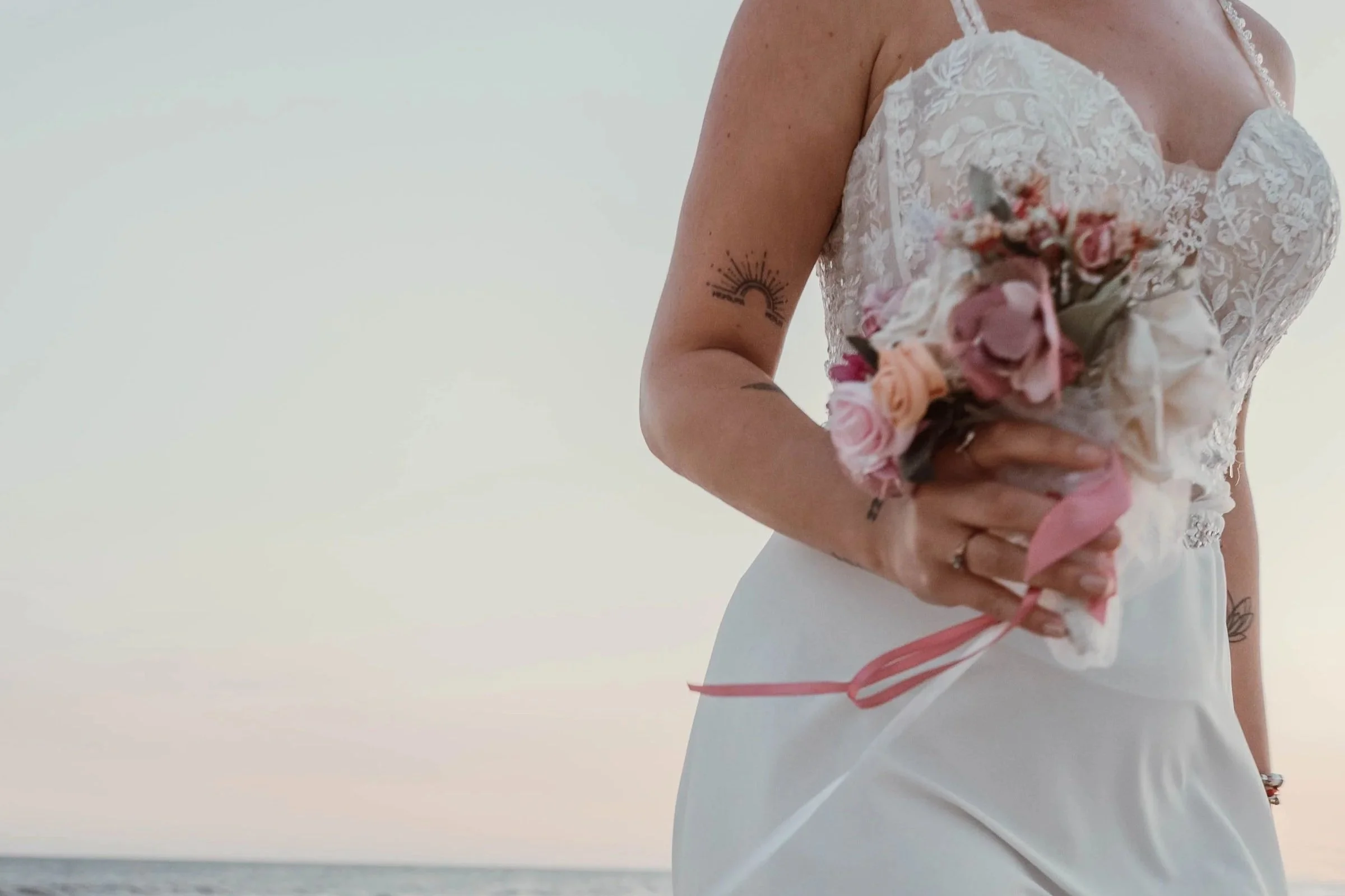 Close-up of a woman in a wedding dress holding a bouquet of pink and peach flowers, standing outdoors with a body of water in the background.