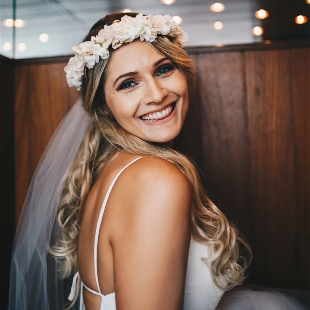 A smiling woman wearing a white dress, floral crown, and veil, posing indoors with warm lighting and wooden wall background.