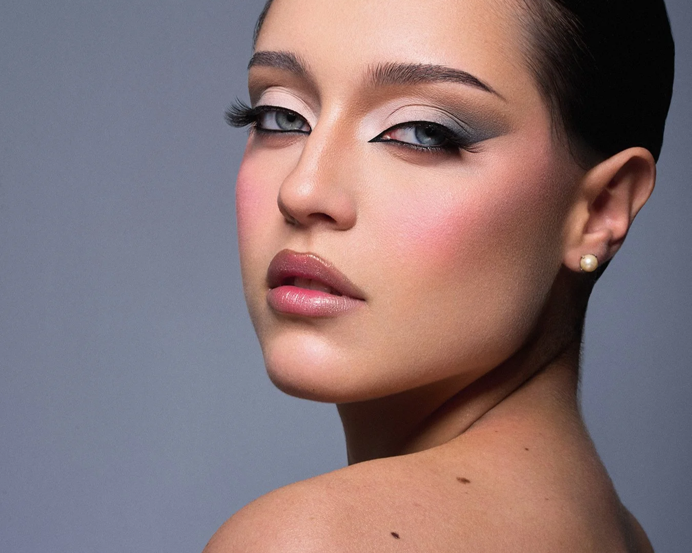 Close-up portrait of a woman with makeup, pearl earring, and dark hair against a gray background.