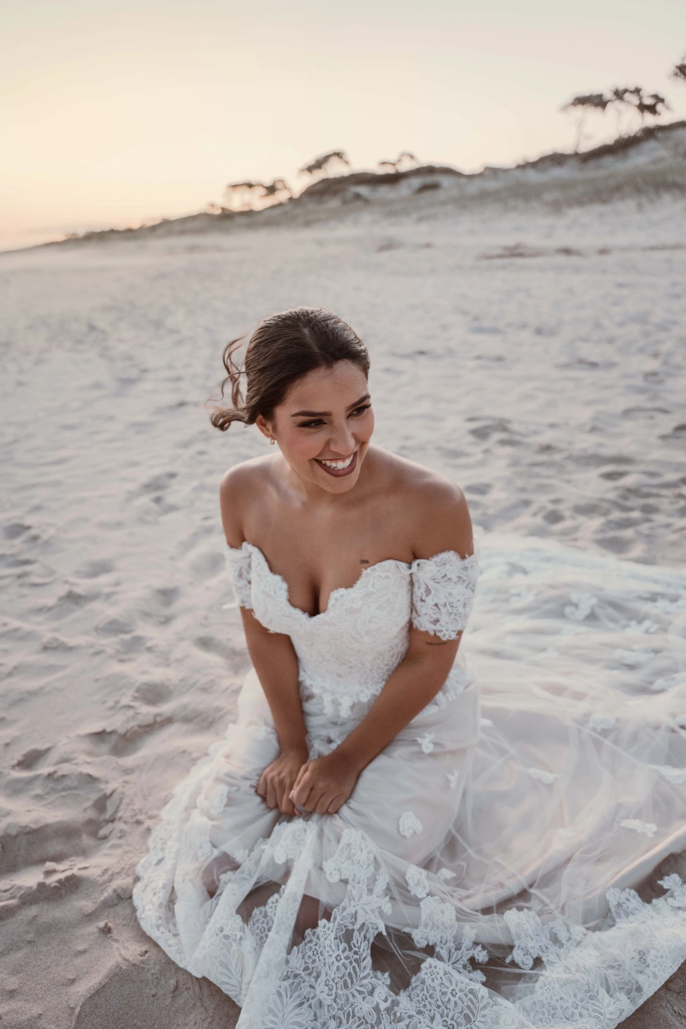 A woman in a white lace wedding dress sitting on the sand at the beach during sunset, smiling and looking to the side.