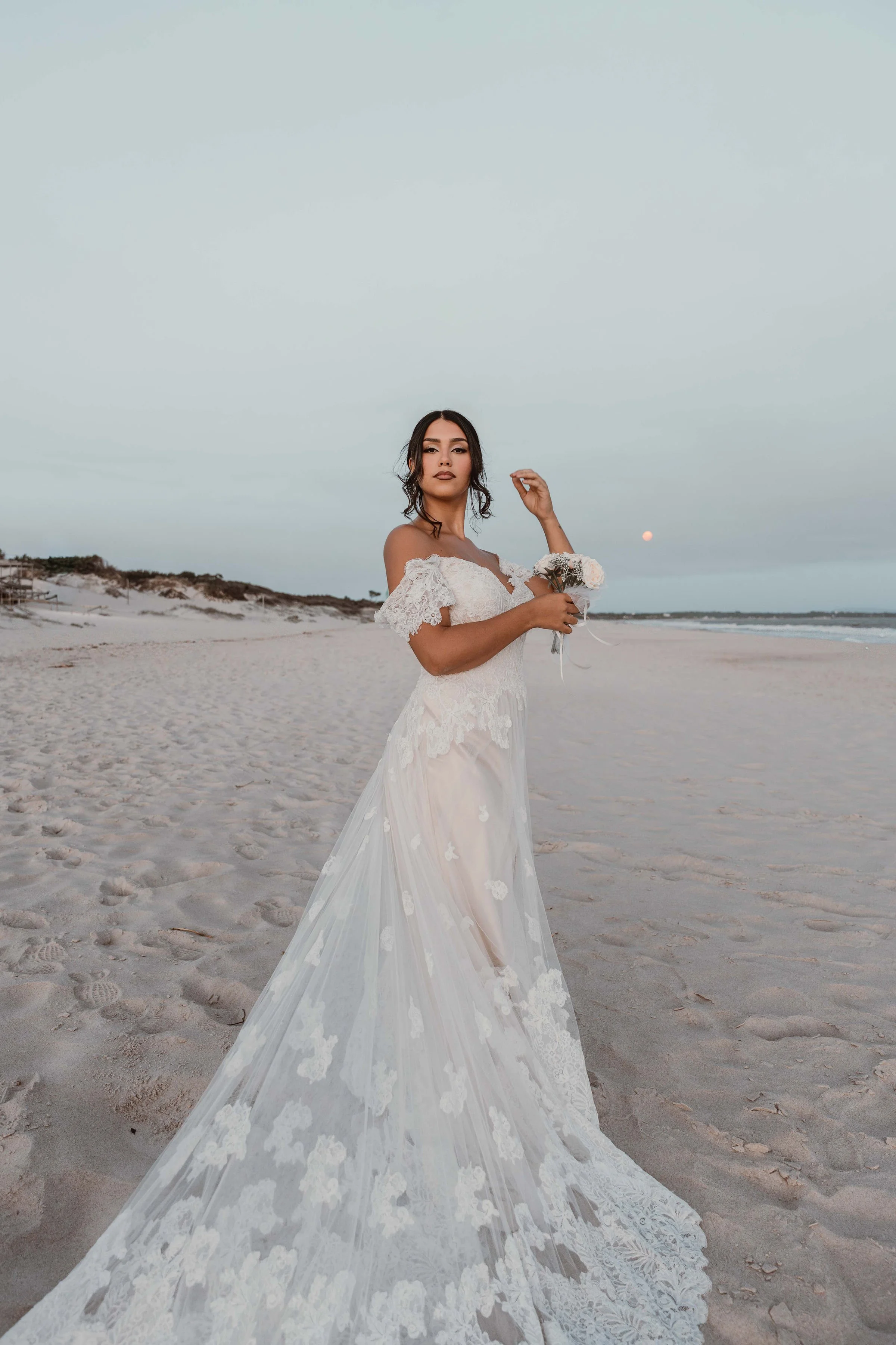 A woman in a white wedding dress holding a small bouquet on a sandy beach at sunset.