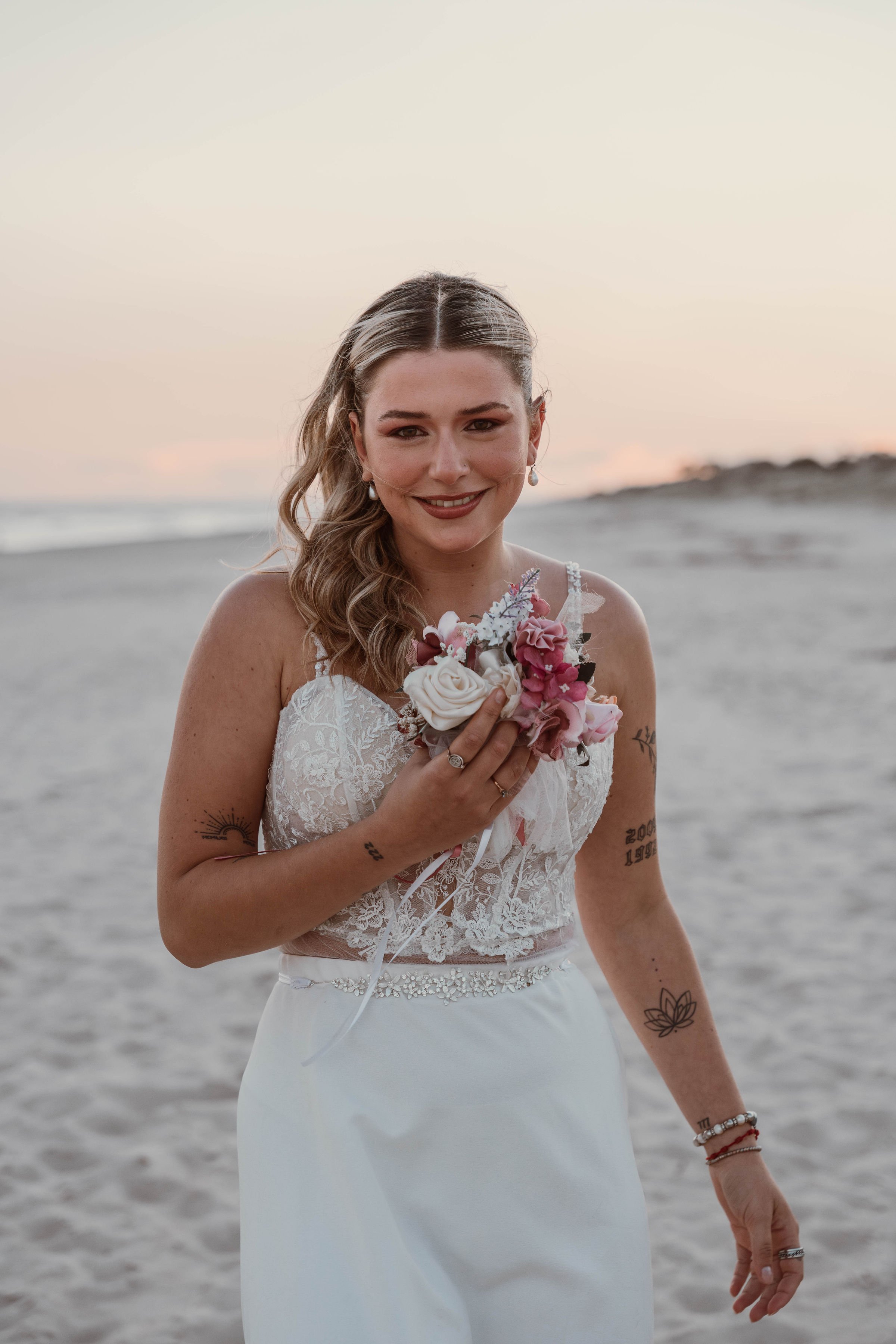 A woman in a wedding dress holding a bouquet of pink and white flowers on a beach at sunset.