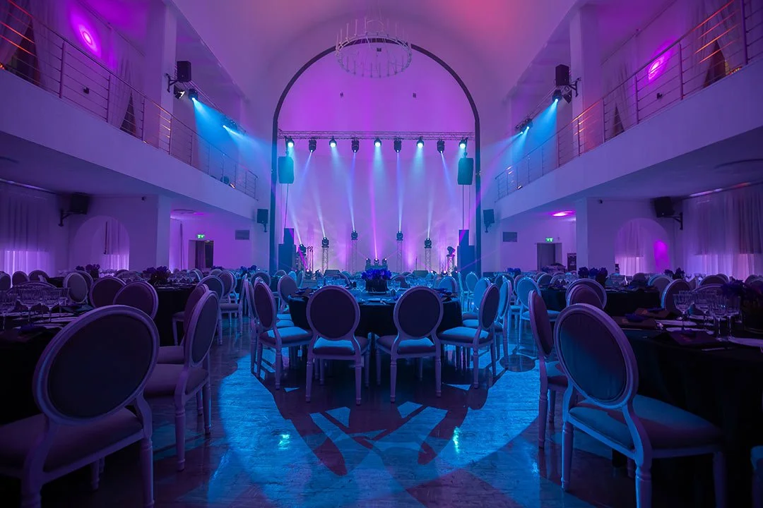 Ein großräumiger festlich dekorierter Saal mit Tischen und Stühlen, beleuchtet in blau- und violettfarbenem Licht, mit einer Bühne und Lichttechnik im Hintergrund, ideal für Veranstaltungen oder Partys.