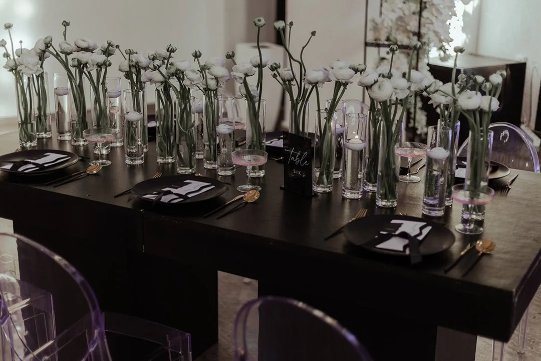Eleganter Esstisch mit weißen Blumen in hohen Glasvasen, schwarzem Geschirr, goldenen Bestecken und Platzkarten, in einem stilvollen Raum