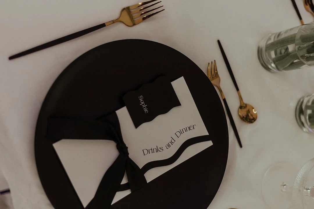 Tischsetup mit schwarzem Teller, schwarze Serviette, schwarze und goldene Besteck, Glasvasen mit Blumen, Menükarte mit 'Drinks and Dinner' und einer schwarzen Namenskarte, alles auf weißem Tischdecken.