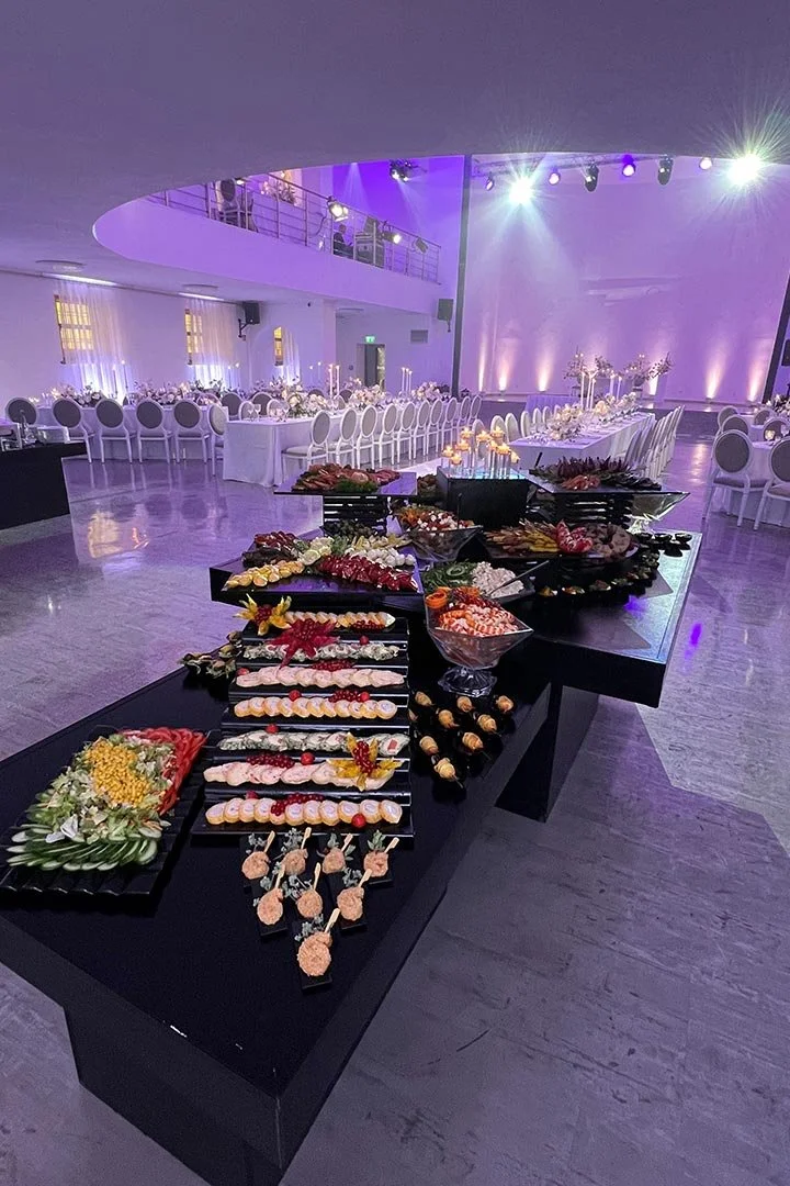 Buffet mit Obst, Canapés und Desserts auf schwarzem Tisch in einem festlich dekorierten Saal mit eleganten Stühlen und Tischen im Hintergrund, blaue und violette Beleuchtung.