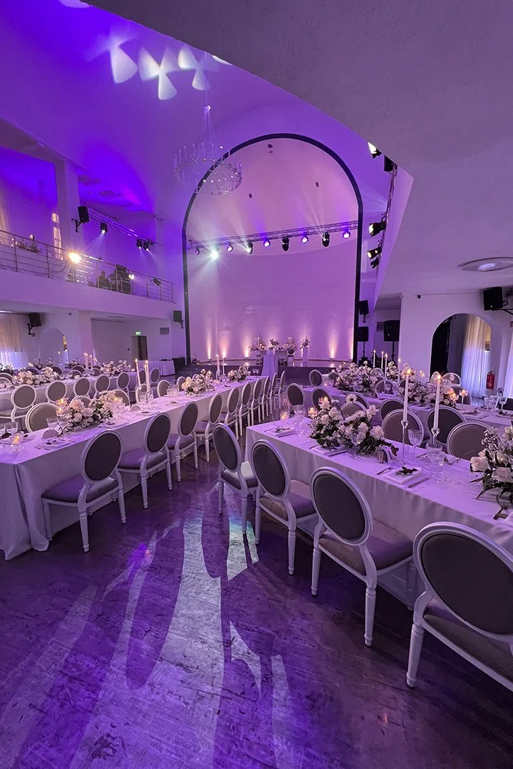 Eleganter Festsaal mit langen Tischen, weißen Tischdecken, Blumenarrangements und Kerzen, ausgestattet für eine Hochzeit oder formelle Veranstaltung, mit blauvioletter Beleuchtung.