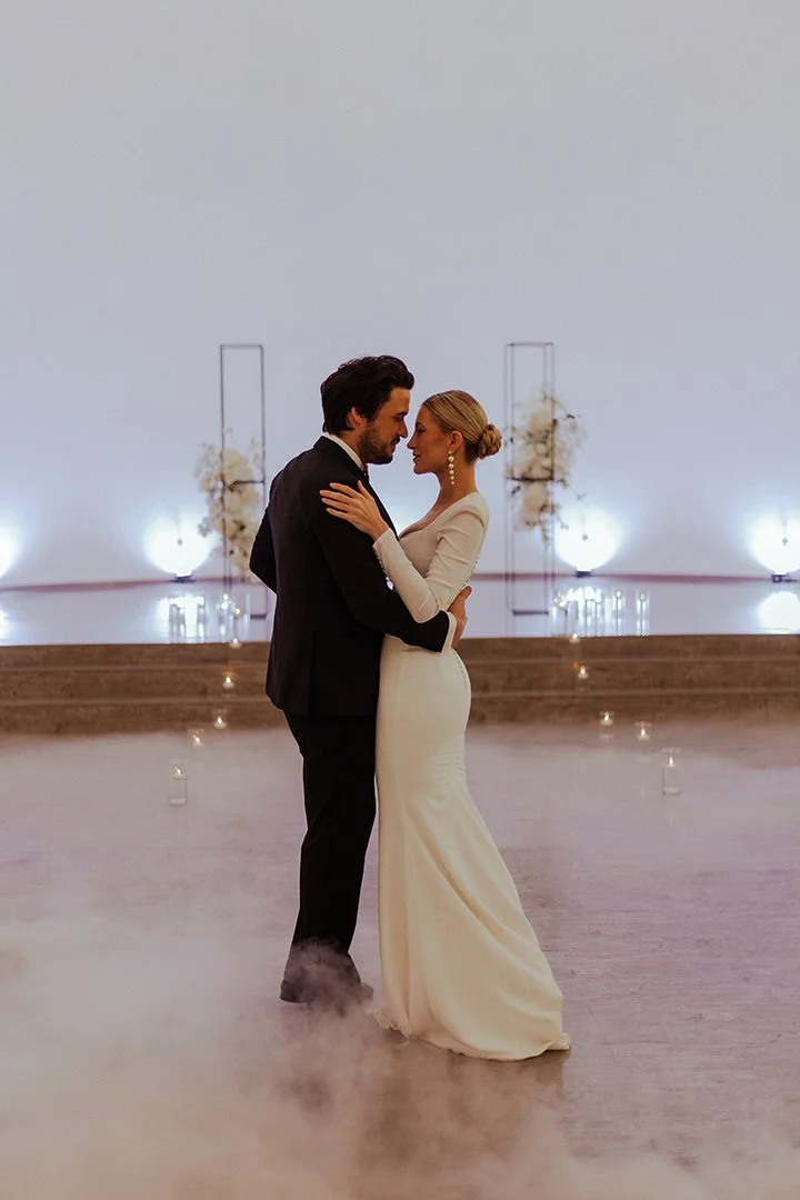 Ein Brautpaar tanzt bei einer Hochzeit in einem eleganten Raum mit Blumenarrangements im Hintergrund.