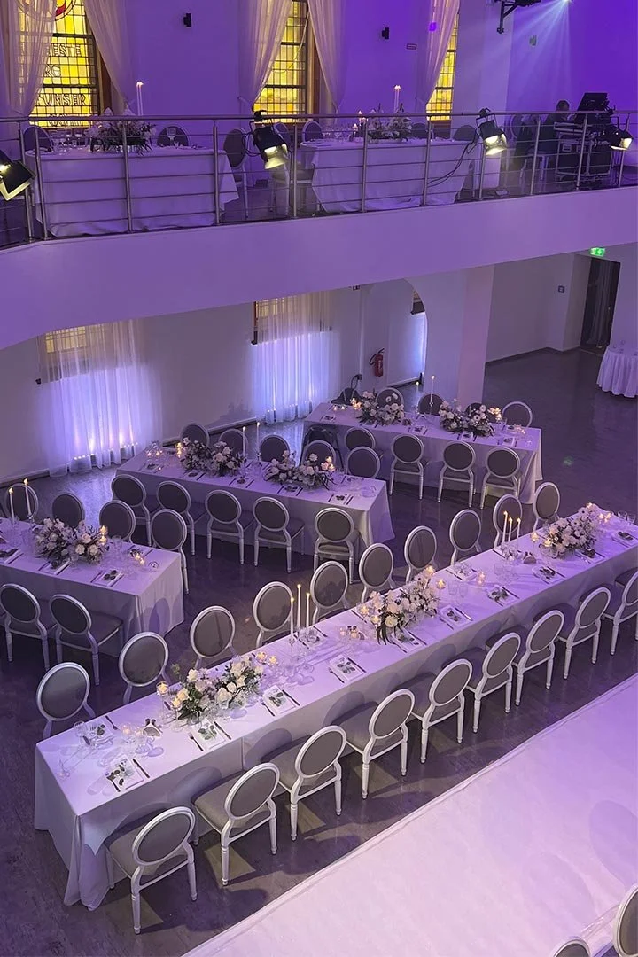 Festlich dekorierter Saal mit langem rechteckigem Tisch und runden Tischen, geschmückt mit Blumen und Kerzen, für eine Feier oder Hochzeit vorbereitet.